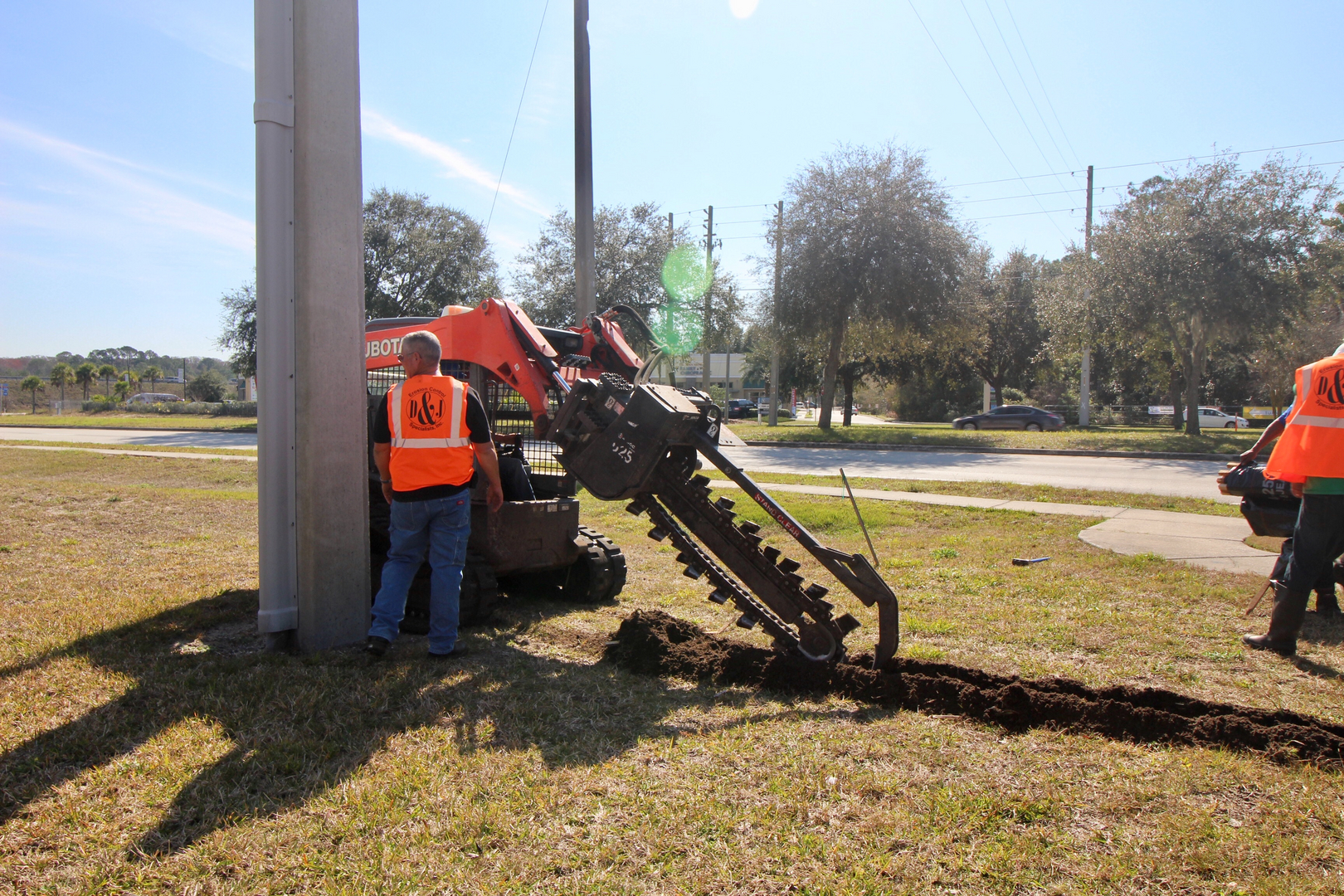 D & J crew operating a skid-steer loader with a trenching attachment beside a utility pole